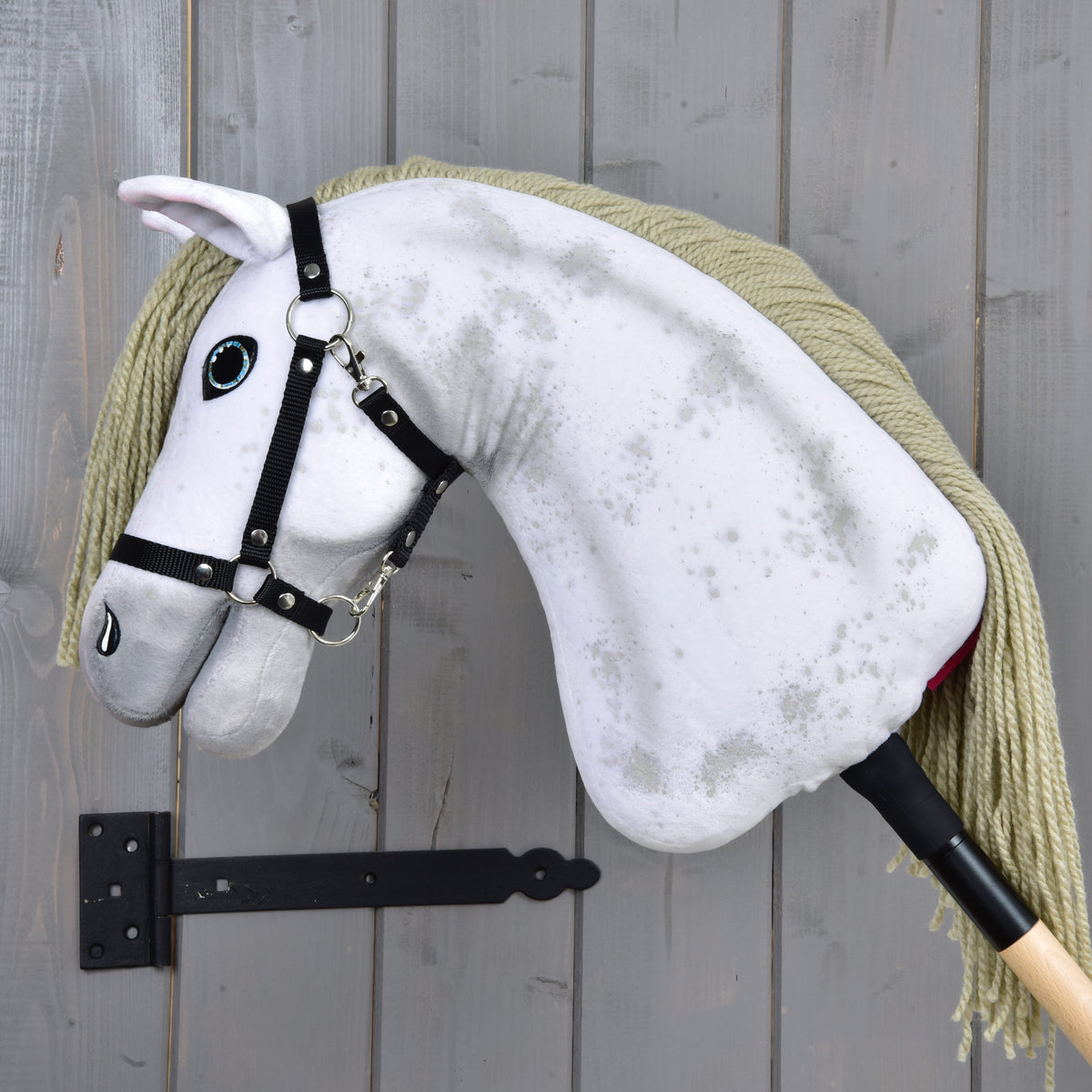 Hobby Horse Victory s čiernou ohlávkou - veľkosť M od Hobby Horse LarDen, s bielym plyšovým prevedením, dlhou hrivou z priadze a čiernou ohlávkou, vystavený na sivej drevenej stene s čiernou kovovou konzolou.