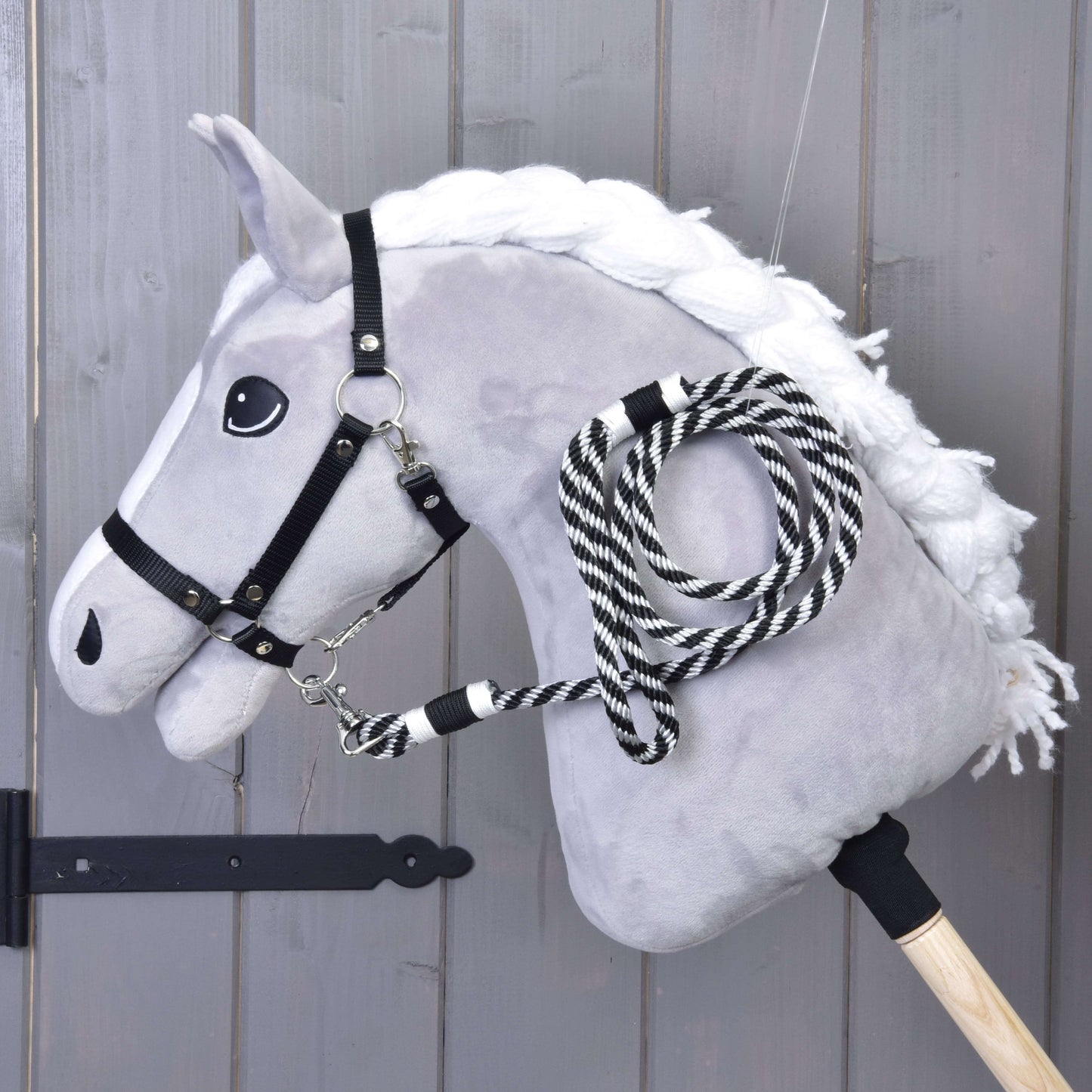 Hobby Horse LarDen „Hobby Horse - vodidlo prúžok čierne“ zobrazuje plyšového koňa s bielou hrivou, čiernou uzdečkou a čiernobielo pruhovaným vodidlom na drevenej tyči, vystaveného pred drevenou stenou.