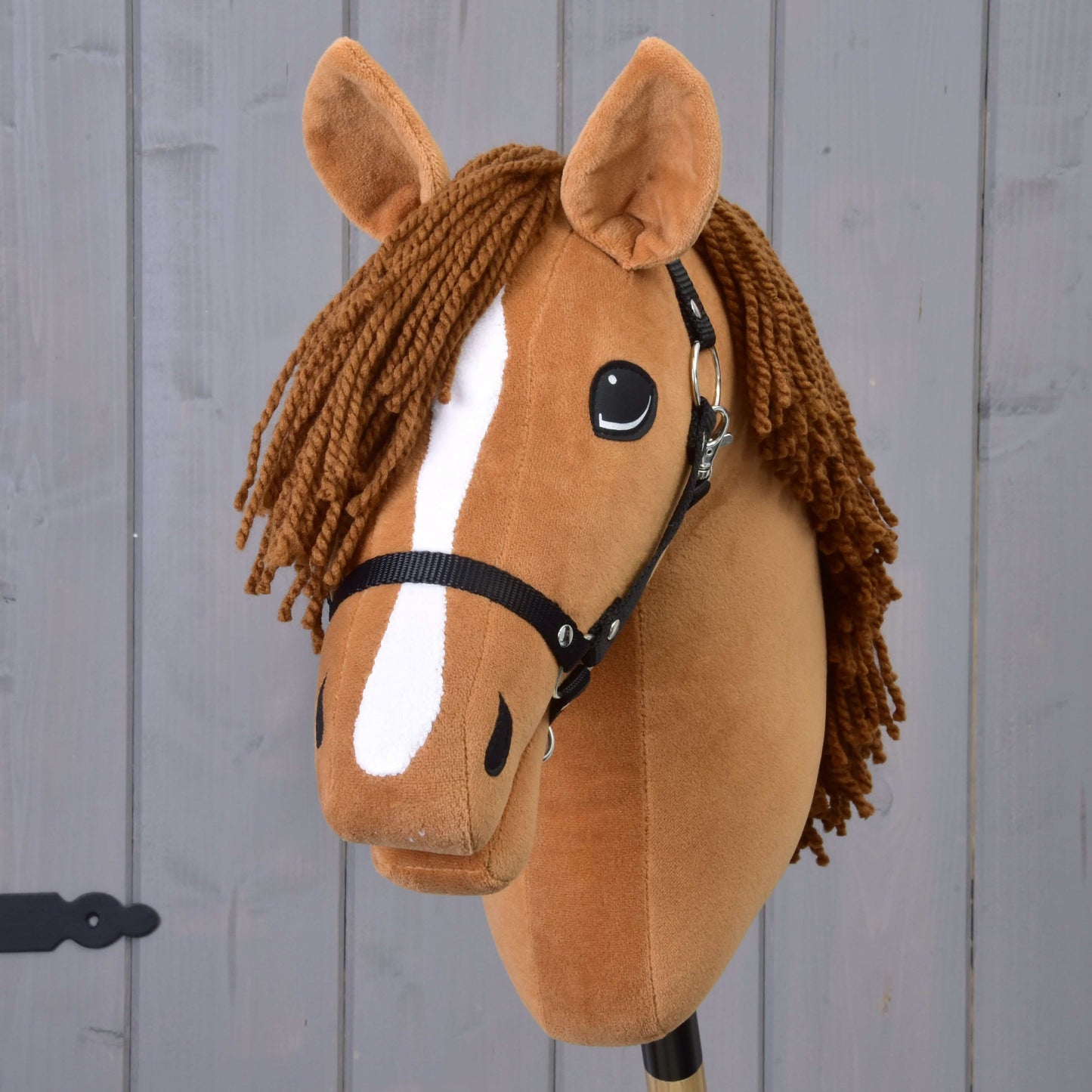 Hobby Horse Wendy s čiernou ohlávkou od Hobby Horse LarDen má hnedú plyšovú hlavu, zapletenú hnedú hrivu z priadze, biely pruh na ňufáku a čiernu ohlávku, inšpirovanú ryšavou kobylou, a je zobrazená na sivom drevenom pozadí.