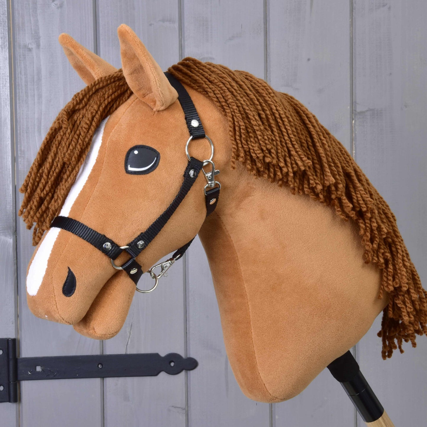 Hobby Horse Wendy s čiernou ohlávkou od Hobby Horse LarDen je plyšová hračka inšpirovaná ryšavou kobylou s hnedou hrivou z priadze, bielym pruhom na tvári a čiernou ohlávkou – ideálna pre nápaditú hru – zobrazená pred dreveným panelovým pozadím.