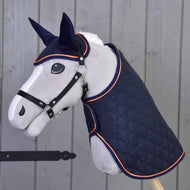 Hobby Horse LarDen "Set Winner" obsahuje vycpanou koňskou hlavu na tyči, oblečenou do prošívané tmavě modré deky a ladící čabraky s oranžovým lemem, naaranžovanou před dřevěnou stěnou.