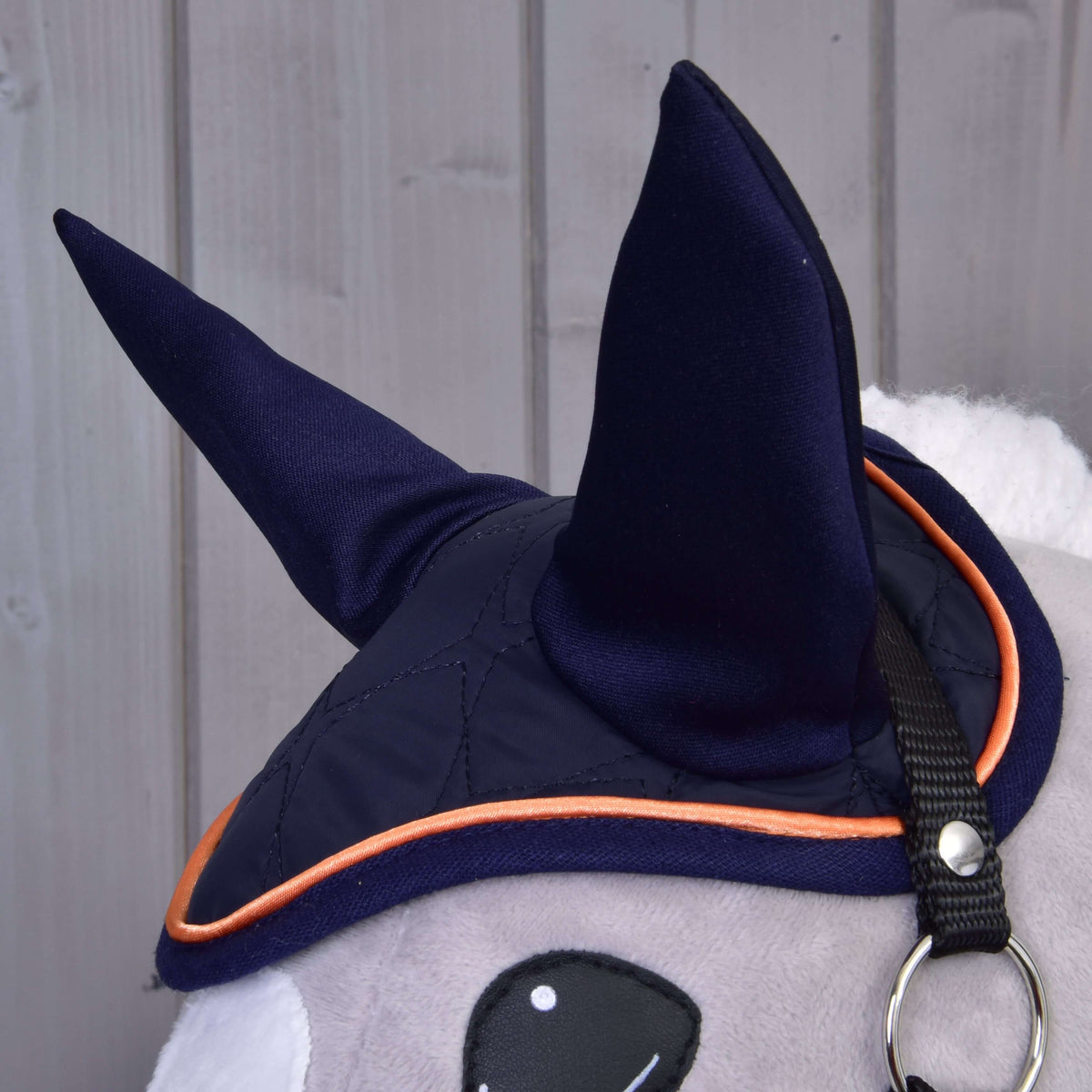 Detail Set Winner od Hobby Horse LarDen zobrazuje plyšové zviera v tmavomodrej čabrake so špicatými ušami a oranžovým lemom na pozadí svetlosivých drevených panelov pre pôvabný vzhľad konskej dekorácie.