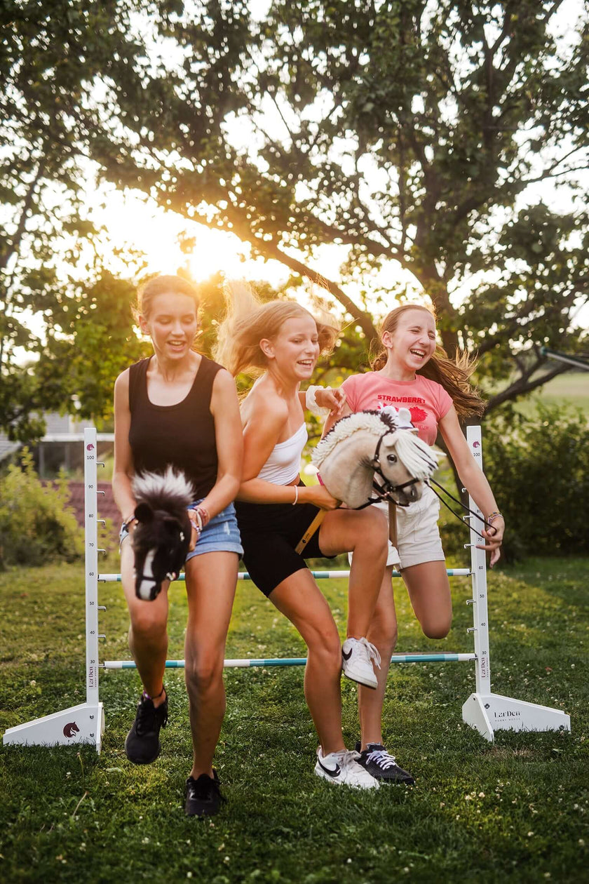 Tri mladé ženy sa smejú, keď vonku bežia a skáču cez prekážku, každá drží Hobby Horse, zatiaľ čo za nimi medzi stromami presvitá slnko a ony si užívajú zábavu pri Hobby Horsingu.