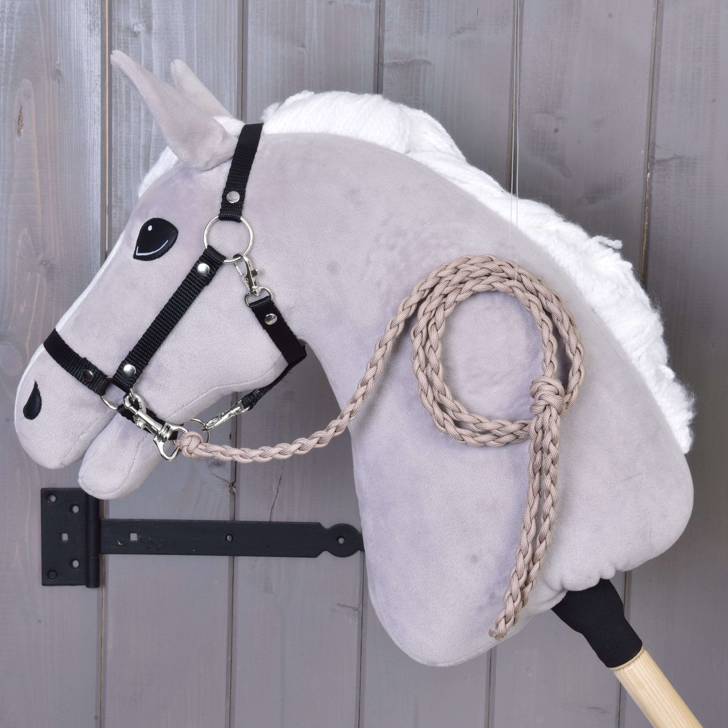 Hobby Horse - pletené vodítko Almond od Hobby Horse LarDen na světle šedém plyšovém Hobby Horse s bílou hřívou, černou uzdečkou a ručně pleteným vodítkem Almond, vystaveném na dřevěné tyči před šedou dřevěnou stěnou.