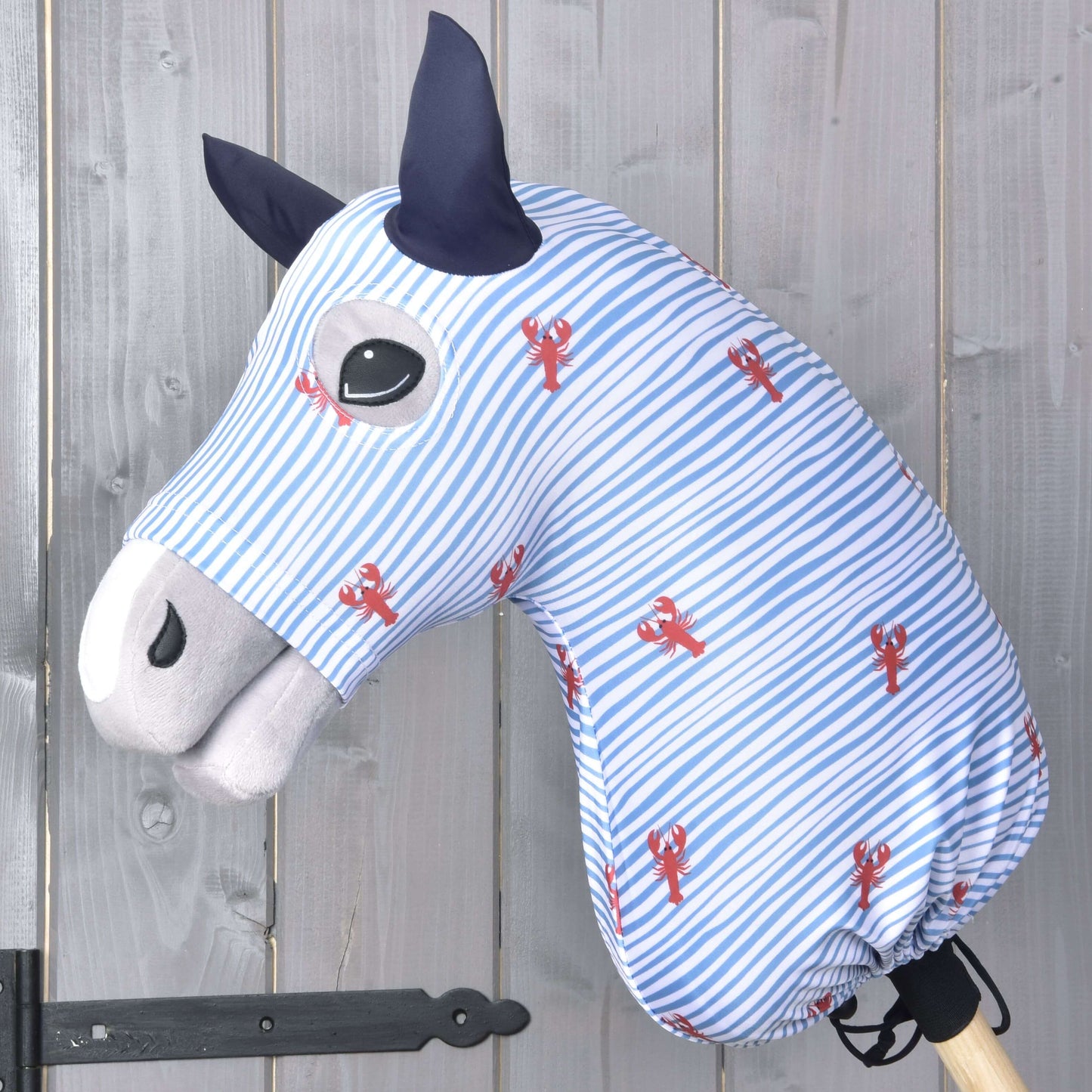 Hobby Horse ochranný oblek Andromeda (Lycra) od Hobby Horse LarDen v modro-bielych pruhoch s červenými potlačami homárov zakrýva hračkárskeho koňa, ktorý má čierne oči, tmavomodré uši a svetlosivý ňufák, na pozadí drevenej steny.