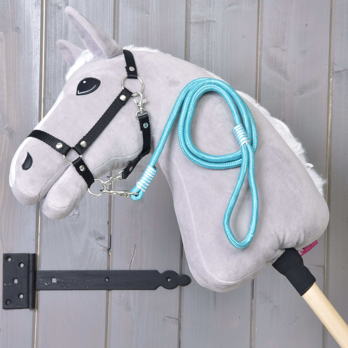 Hobby Horse - vodidlo Atlantis od Hobby Horse LarDen so sivou plyšovou hlavou, čiernou uzdečkou a modrým povrazovým vodidlom, upevnené na drevenej tyči a zobrazené pred zvislými drevenými doskami.