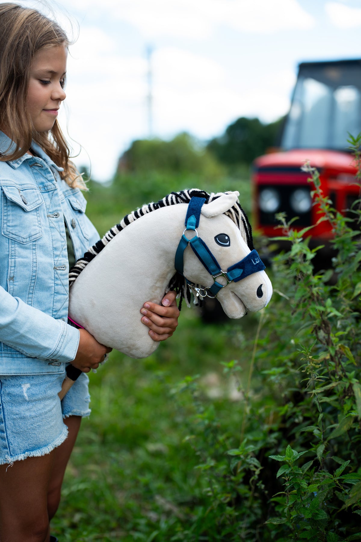 Dievča v džínsovej bunde drží Hobby Horse Aurora s čiernou ohlávkou od LarDen pri živom plote, v pozadí rozmazaný červený traktor, krémovobiela plyšová hlava a čiernobiela pruhovaná priadzová hriva, s ohlávkou LarDen Blue s kožušinou