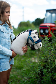 Dievča v džínsovej bunde drží Hobby Horse Aurora s čiernou ohlávkou od LarDen pri živom plote, v pozadí rozmazaný červený traktor, krémovobiela plyšová hlava a čiernobiela pruhovaná priadzová hriva, s ohlávkou LarDen Blue s kožušinou