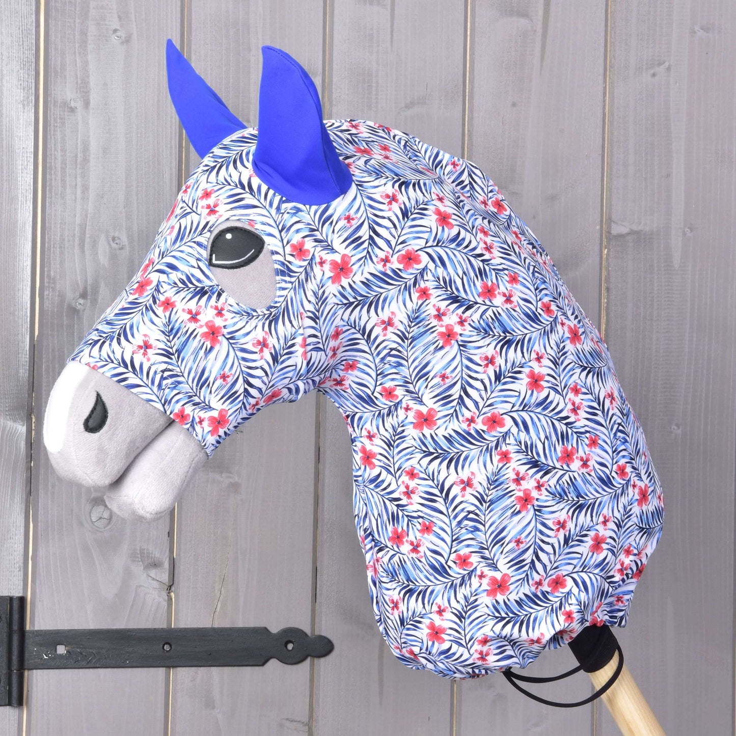 Hobby Horse LarDen Hobby Horse ochranný oblek Avatar (Lycra) s látkovou hlavou s modrým a červeným květinovým vzorem, modrými plstěnými ušima, šedou hřívou a pratelným obalem. Je nasazený na dřevěné tyči před dřevěným panelovým pozadím.