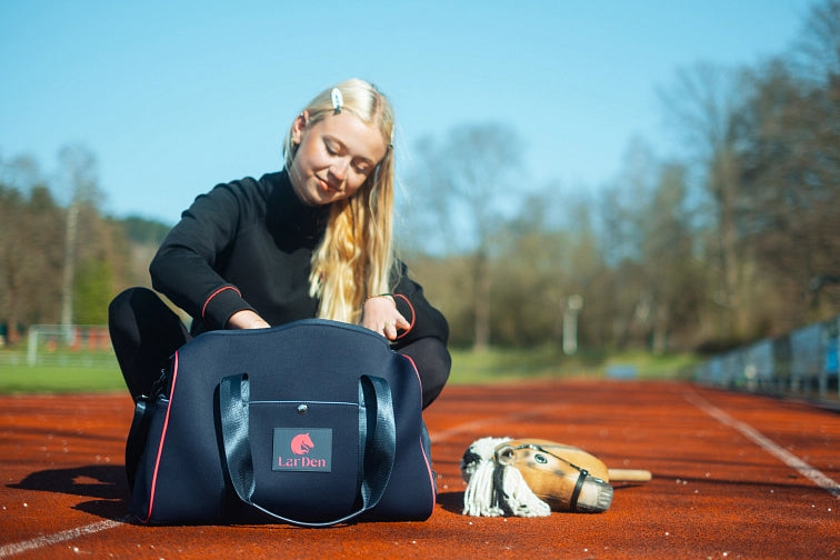 Mladá žena s dlhými blond vlasmi sedí na atletickej dráhe a pozerá sa do tmavomodrej športovej tašky s nápisom LarDen. Vedľa nej na zemi leží Hobby Horse, naznačujúci jej vášeň pre Hobby Horsing. V pozadí vidno stromy a čiary atletickej dráhy.