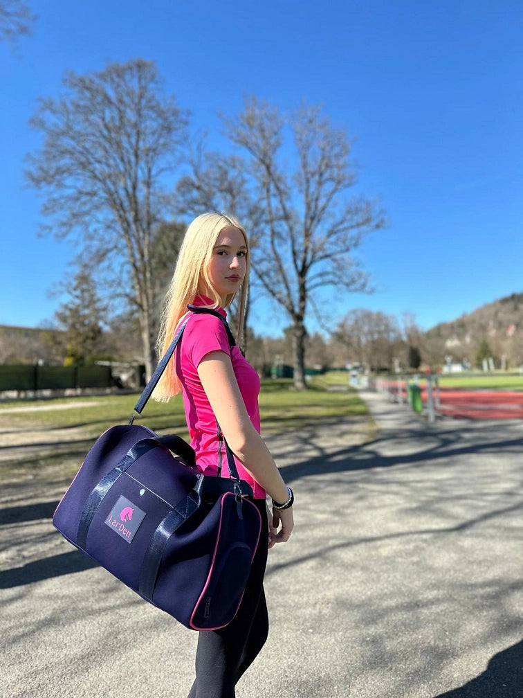 Mladá žena s dlhými blond vlasmi v ružovom športovom tričku a s tmavomodrou športovou taškou stojí vonku za slnečného dňa so svojím Hobby Horse, pripravená na zábavný deň Hobby Horsingu pod jasne modrou oblohou a medzi zelenými stromami.