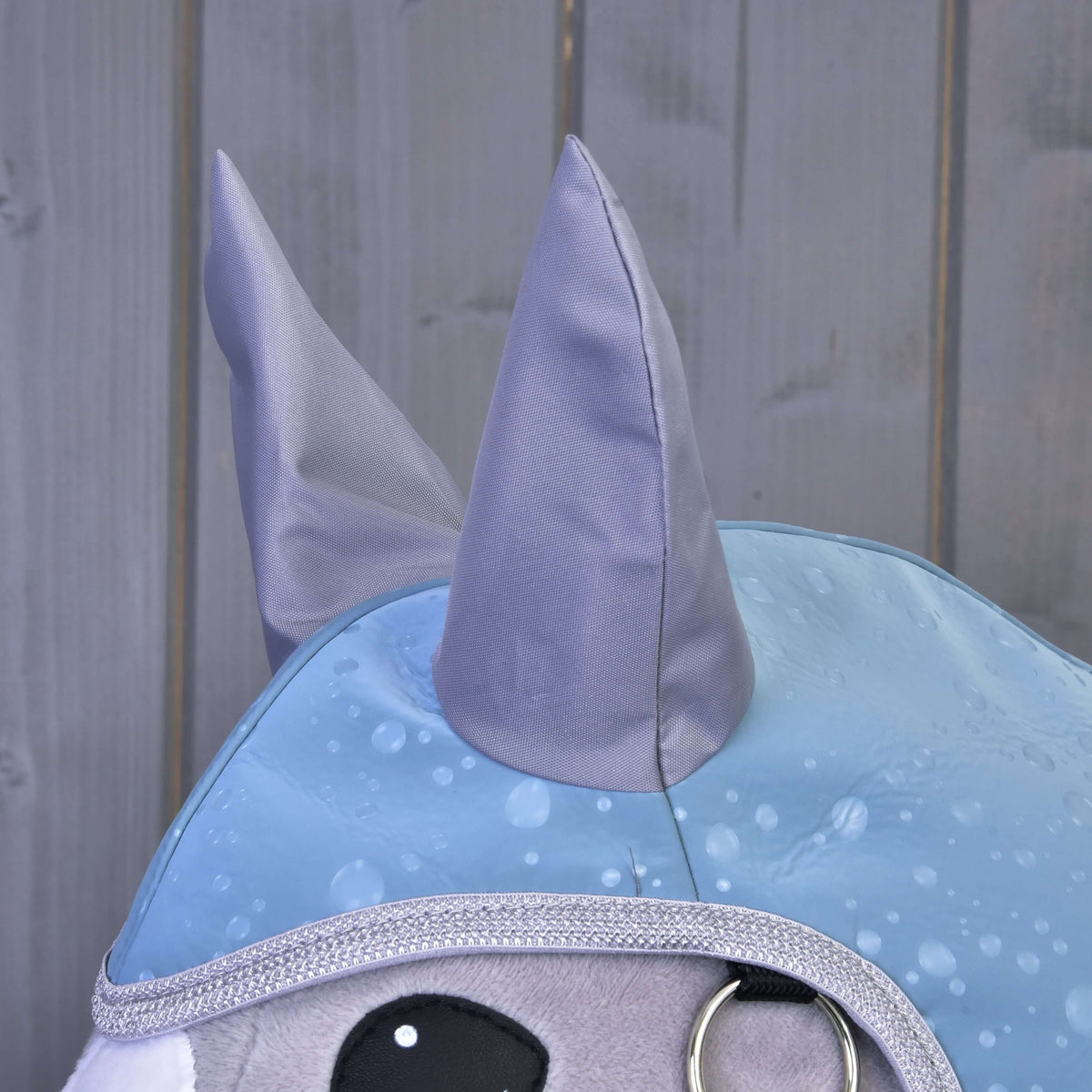 Detail plyšovej hračky Hobby Horse LarDen v outfite „Pláštenka Blue Drops“ s lesklými sivými ušami, strieborným lemovaním, veľkými čiernymi očami a modrým plášťom so vzorom kvapiek dažďa. V pozadí sú hladké sivé drevené panely.