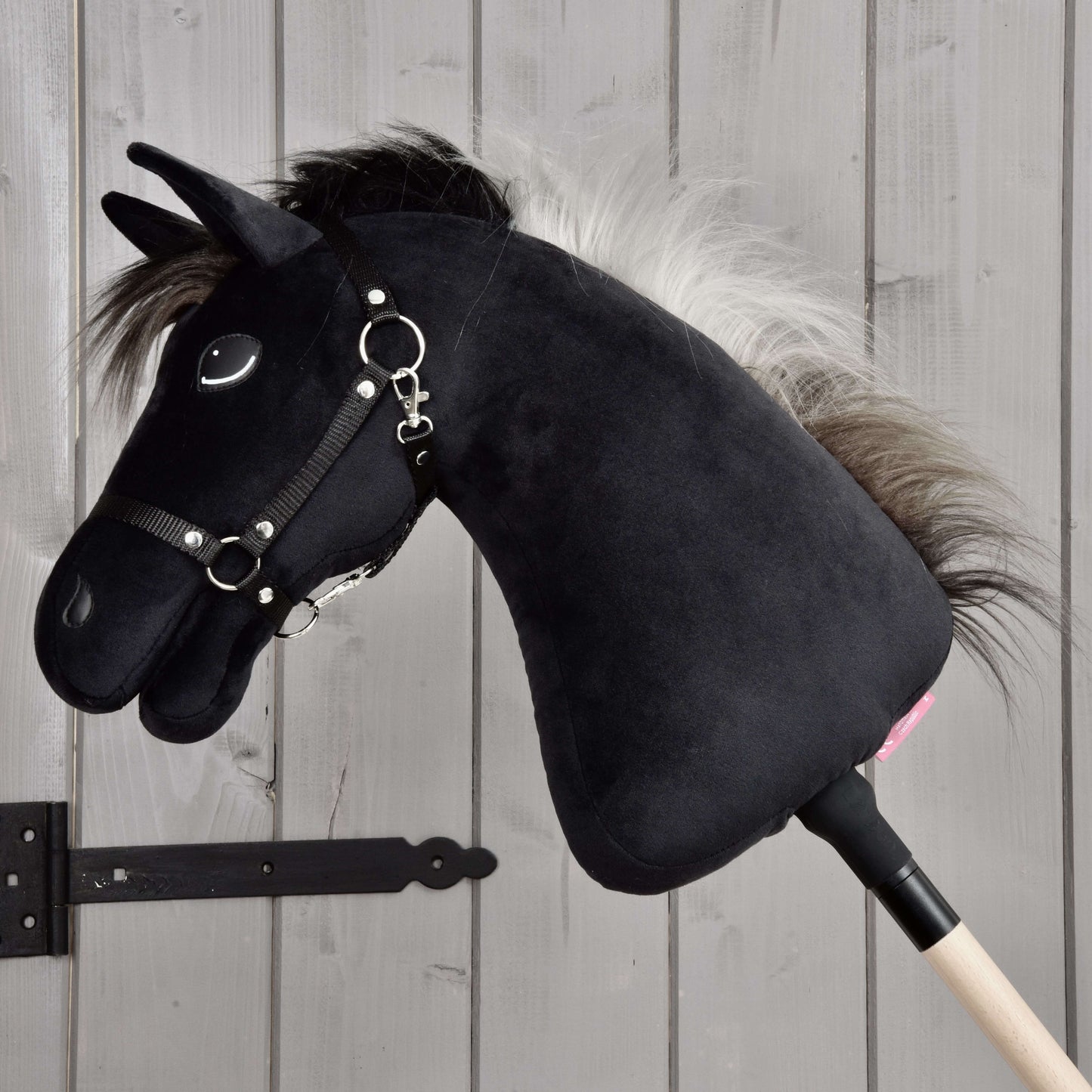 Hobby Horse Bond s čiernou ohlávkou od Hobby Horse LarDen, čierny plyšový Hobby Horse s bielou hrivou, čiernou ohlávkou a opratami na drevenej tyči, zobrazený pred svetlou drevenou stenou – ideálny pre každého fanúšika Hobby Horse Bond.