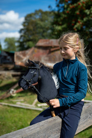 Blond dievča v petrolejovom tope sedí na drevenom zábradlí na slnečnom dvore a drží Hobby Horse Bond s čiernou ohlávkou od LarDen, čierna plyšová hlava a tmavohnedá priadzová hriva, s ozdobnou bezudidlovou uzdečkou