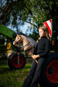 Dievča v čiernom roláku stojí pri zelenom poľnohospodárskom stroji a drží Hobby Horse Bren s čiernou ohlávkou od LarDen, pieskovohnedá plyšová hlava a krémová priadzová hriva, s náprsenkou Espresso s kožušinou