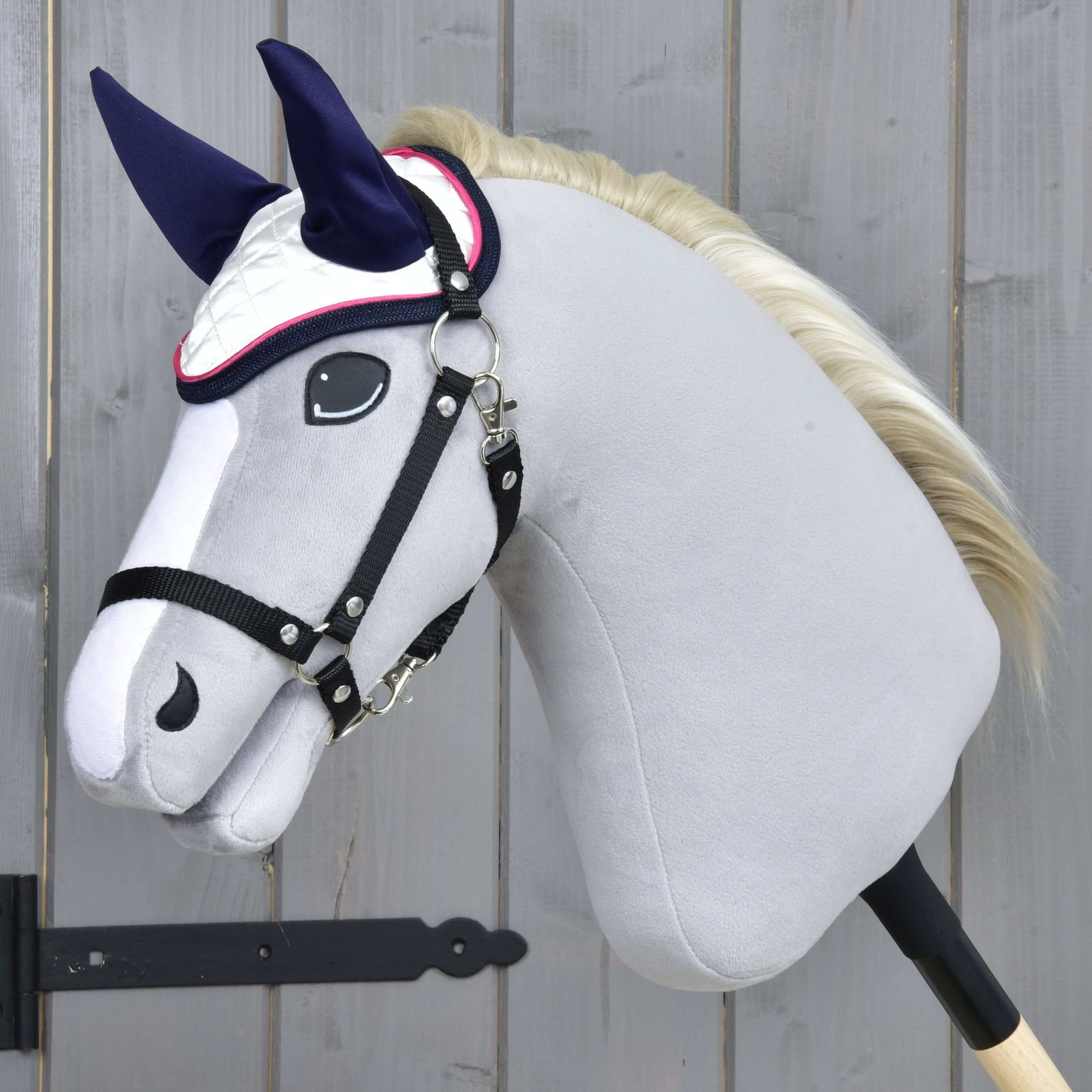 Čabraka Number 1 od Hobby Horse LarDen na ručne vyrábanom sivom Hobby Horse – biela s tmavomodrými ušami na ochranu proti hmyzu – s bielou hrivou, čiernou uzdečkou, nasadenom na drevenej tyči pred pozadím zo svetlých drevených panelov.
