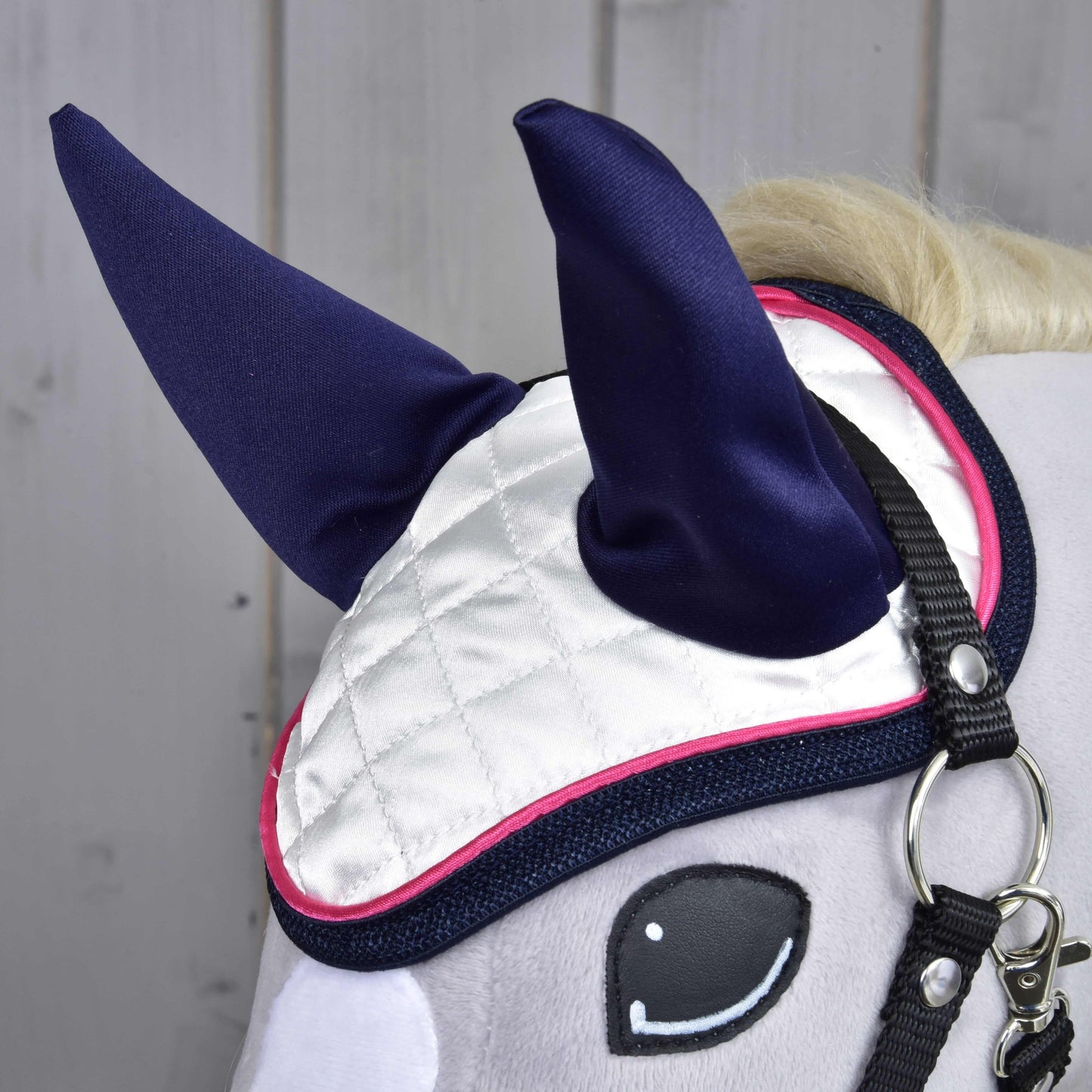 Detail hlavy Hobby Horse LarDen s čabrakou Number 1 – biela prešívaná s tmavomodrými ušami a ružovým lemovaním – na ochranu proti hmyzu, doplnená čiernou uzdečkou, na svetlom drevenom pozadí.