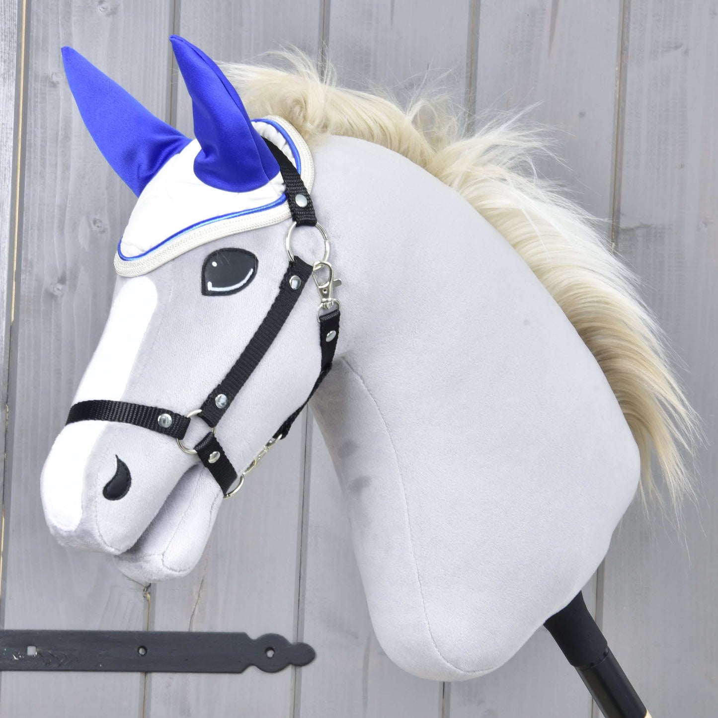 Hobby Horse LarDen se světle šedou plyšovou hlavou, bílým čenichem, černou uzdečkou a krémově zbarvenou hřívou, doplněný o čabraku Cream Pearl k ochraně proti hmyzu. Je nasazený na tyči a umístěný na dřevěném pozadí.