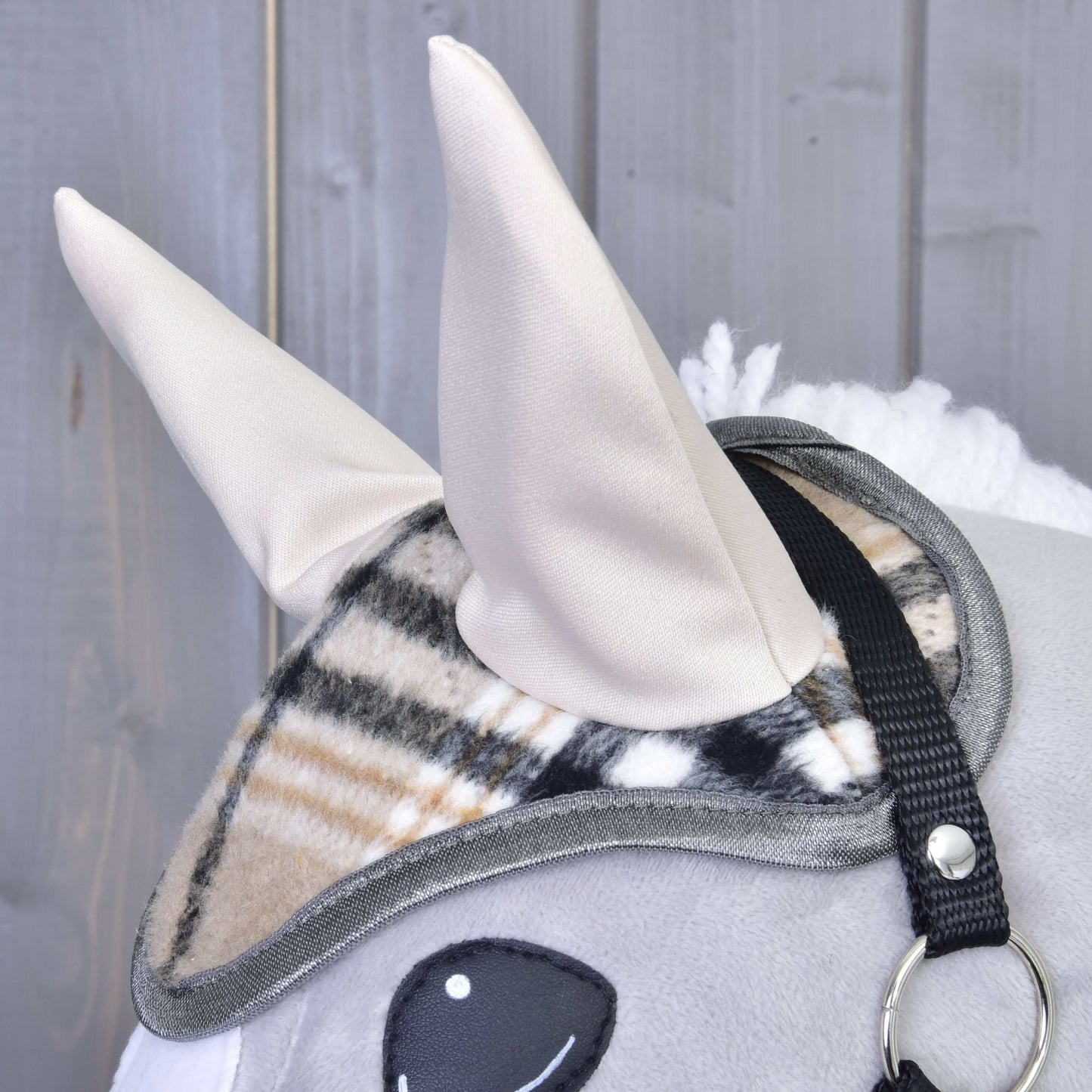 Detail plyšového Hobby Horse od Hobby Horse LarDen s dvoma béžovými látkovými ušami, s čabrakou Cremo England s károvaným vzorom na ochranu pred muchami a čiernym remienkom uzdečky, na sivom drevenom pozadí.