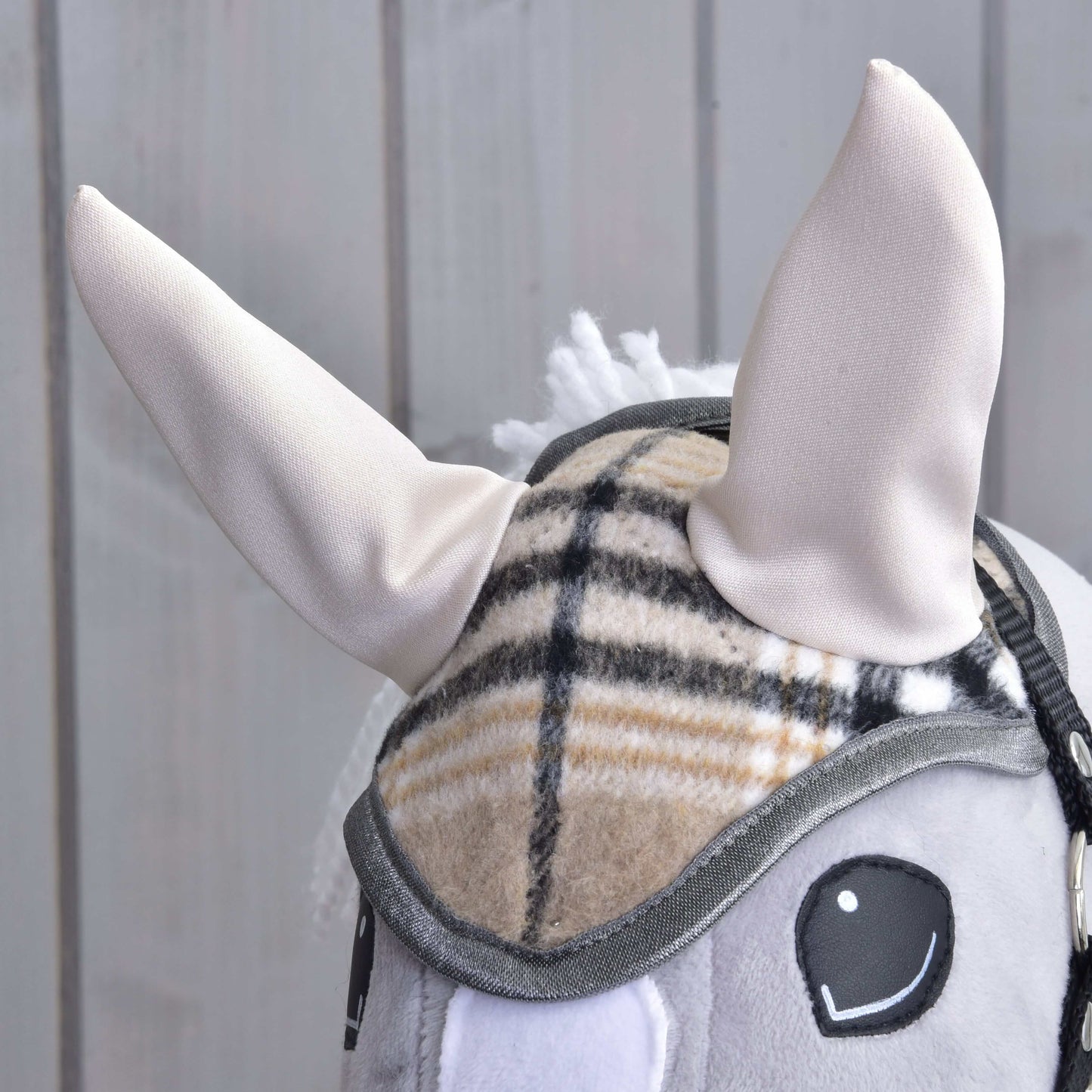 Detail Hobby Horse LarDen Čabraka Cremo England: má mäkké krémovo sfarbené uši, károvaný remienok uzdečky na ochranu pred muchami a sivú hrivu, zobrazená na svetlom drevenom pozadí.