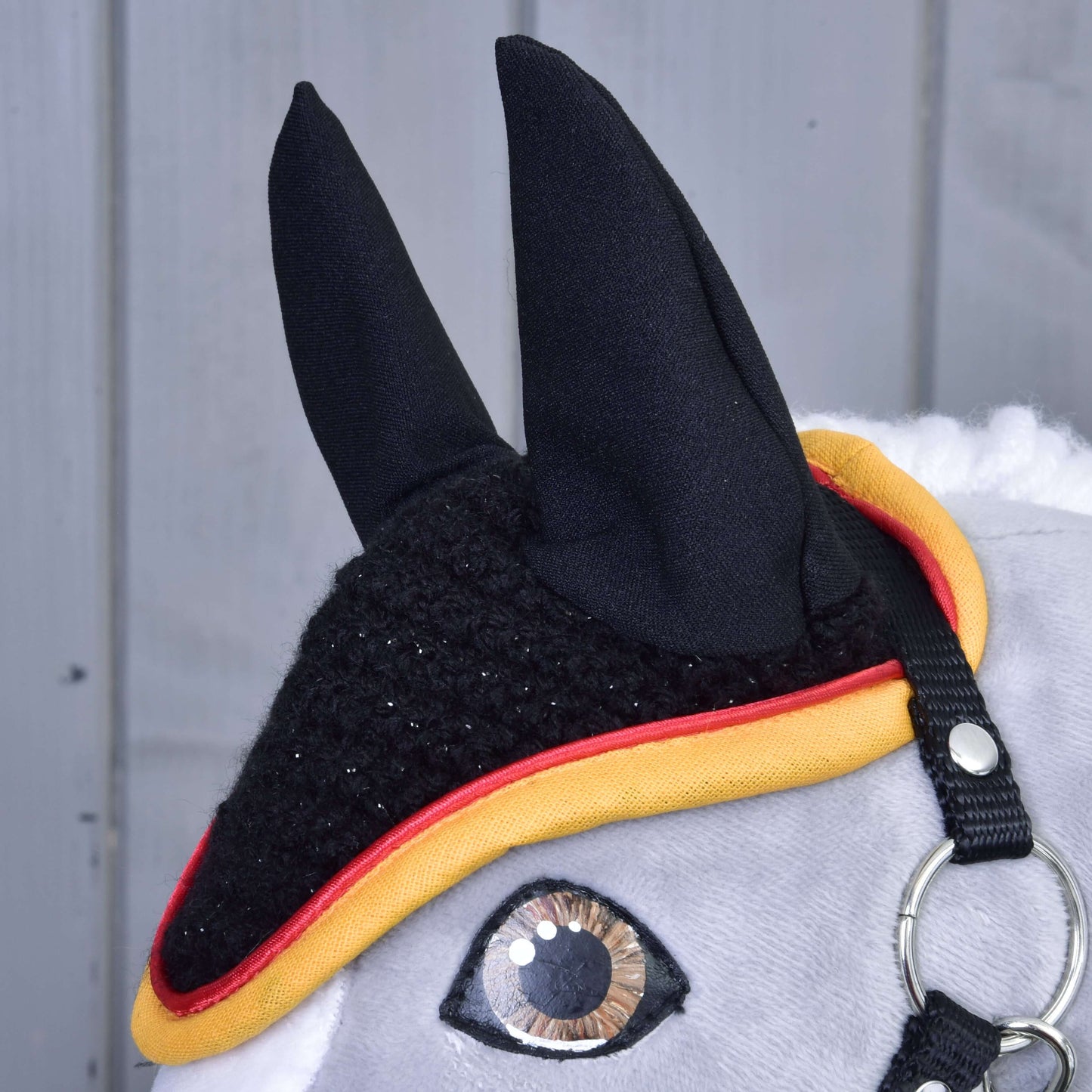 Detail šedého Hobby Horse od Hobby Horse LarDen s „Čabraka háčkovaná Flag“ pro ochranu proti hmyzu, se žlutým a červeným lemováním, jedním lesklým okem, kovovým kroužkem uzdečky a jemnou hřívou na pozadí šedého dřeva.