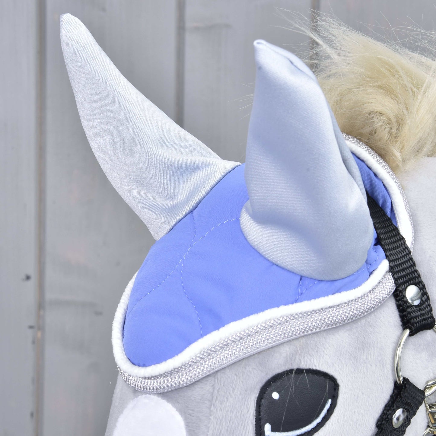 Detail světle šedé plyšové hračky Hobby Horse od Hobby Horse LarDen s čabrakou Light Violett na ochranu proti mouchám, s lesklým lemováním, černými řemínky postroje a bílým a černým okem na dřevěném pozadí.