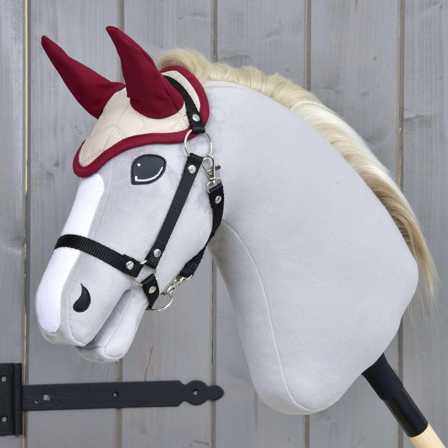 Čabraka Maroon Star od Hobby Horse LarDen na plyšovom Hobby Horse s bielym telom, čiernou uzdečkou a krémovou hrivou, nasadenom na tyči na drevenom pozadí, na ochranu proti hmyzu.