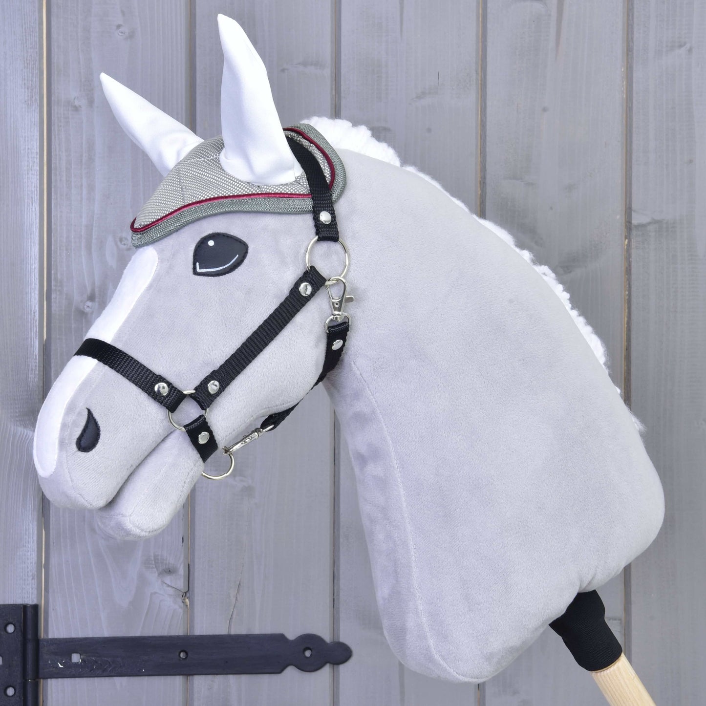Detail sivého plyšového Hobby Horse od Hobby Horse LarDen s uzdečkou, ozdobným čelenkovým remienkom a čabrakou Silver Eleganza na ochranu pred hmyzom, na pozadí dreveného plota.