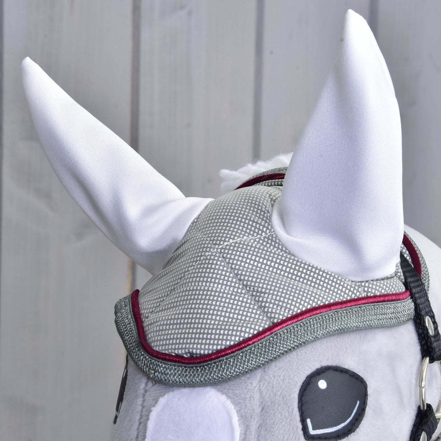 Detail Hobby Horse LarDen Čabraka Silver Eleganza: sivá sieťovaná čabraka s červeným lemovaním, veľkými vztýčenými bielymi ušami a čierne vyšívanými očami, ideálna na ochranu pred hmyzom, na svetlom drevenom pozadí.