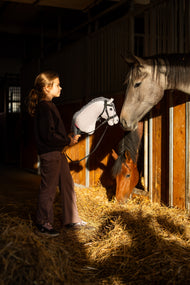 Tmavovlasé dievča v čiernej mikine stojí v uličke stajne s Hobby Horse Caesar s čiernou ohlávkou od Hobby Horse LarDen, sivou plyšovou hlavou s čierno-bielou hrivou z priadze, kým sivý a hnedý kôň pozerajú z boxov, s ozdobenou kompletnou bezudidlovou uzdečkou