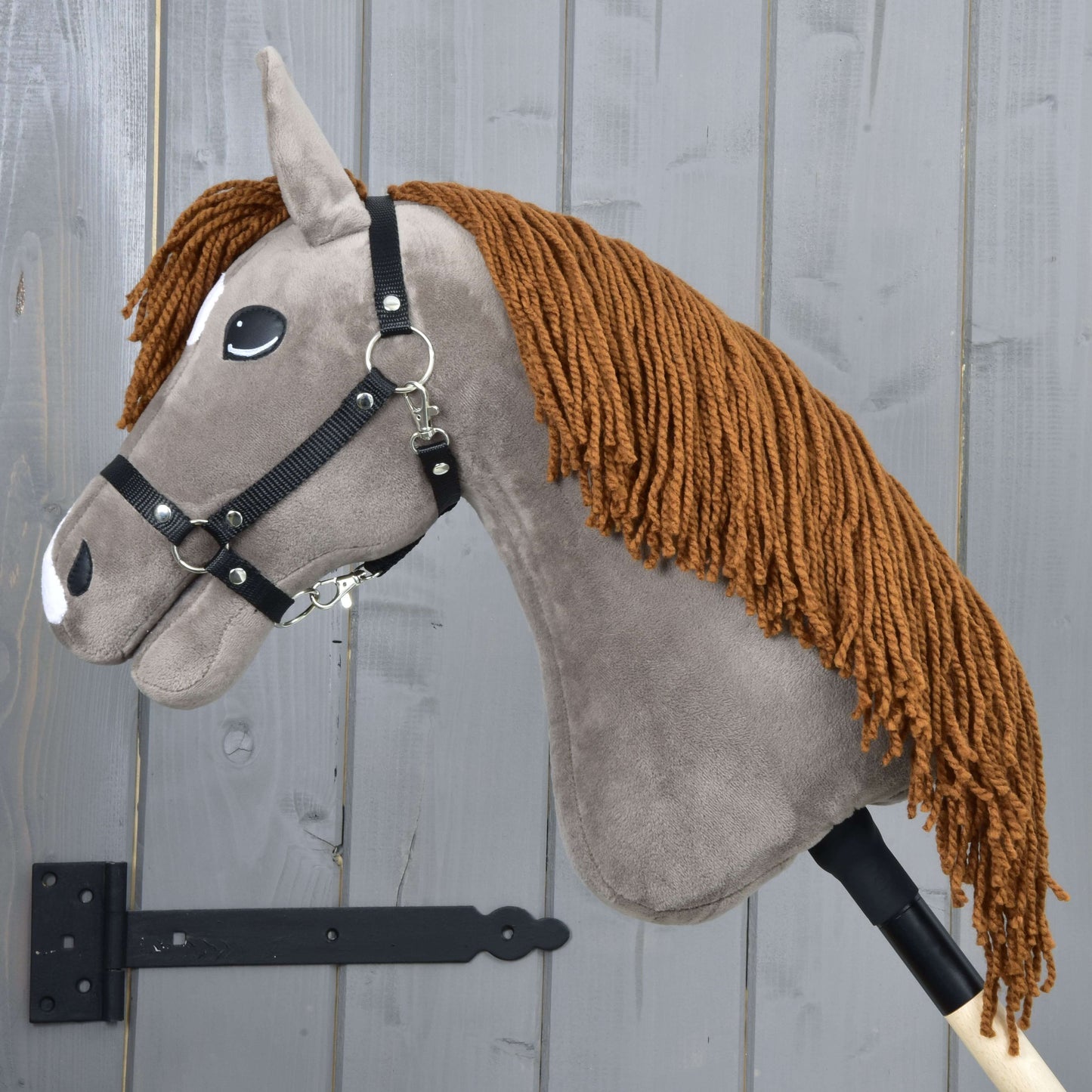 Hobby Horse Chanell s čiernou ohlávkou od Hobby Horse LarDen, so sivou látkovou hlavou, hnedou priadzovou hrivou, čiernou ohlávkou a bielymi plstenými detailmi, nasadený na drevenej tyči pred stenou zo sivých drevených dosiek.