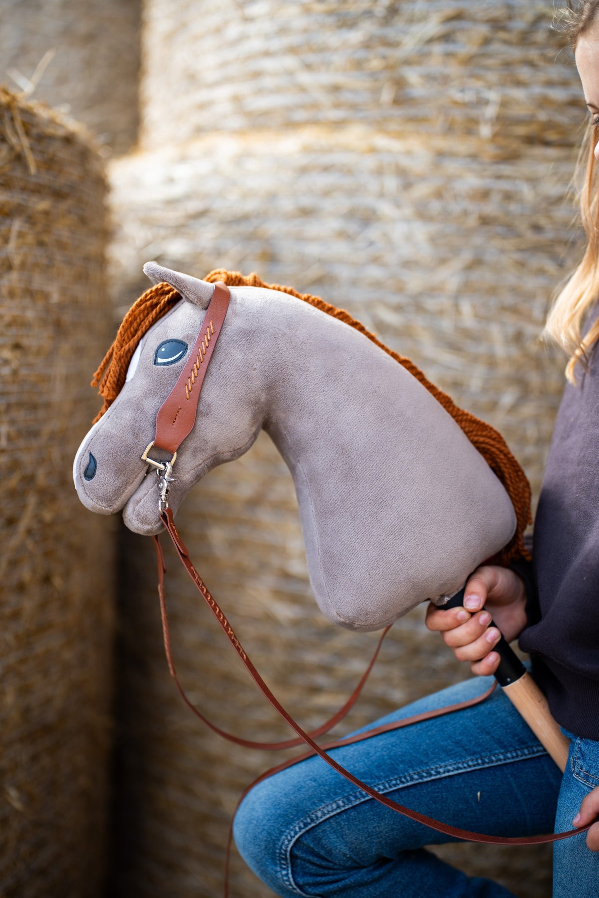 Hnedá jednoušná uzdečka s oťažami na sivom plyšovom Hobby Horse so zázvorovou priadzovou hrivou pri balíkoch slamy