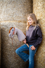 Usmievajúce sa dievča v tmavohnedej mikine sa opiera o okrúhle balíky slamy a drží Hobby Horse Chanell with Black Halter od Hobby Horse LarDen so sivou plyšovou hlavou, modrým okom a hrdzavou priadzovou hrivou, s kompletnou westernovou uzdečkou s jedným náušníkom