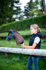 Dievča v tmavomodrej prešívanej veste stojí za bielym ohradníkom a drží Hobby Horse Chianti with Black Halter od Hobby Horse LarDen, za ním je strihaný zelený živý plot, s tmavohnedou plyšovou hlavou a tmavou priadzovou hrivou, s ohlávkou Teddy Petrol s kožušinovou podšívkou