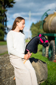 Dievča v krémovom svetri sedí na balvane pri lúčnej ceste a drží Hobby Horse Cloony with Black Halter od Hobby Horse LarDen s čiernou plyšovou hlavou, bielou hviezdou a čiernou priadzovou hrivou, s čiernou koženkovou ohlávkou s kompletnou ružovou kožušinou