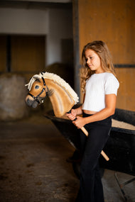 Dievča v bielom tričku stojí v stajni a drží Hobby Horse Colby with black Halter od Hobby Horse LarDen s palomino plyšovou hlavou, hustou krémovou priadzovou hrivou a čiernou ohlávkou so strieborným kovaním