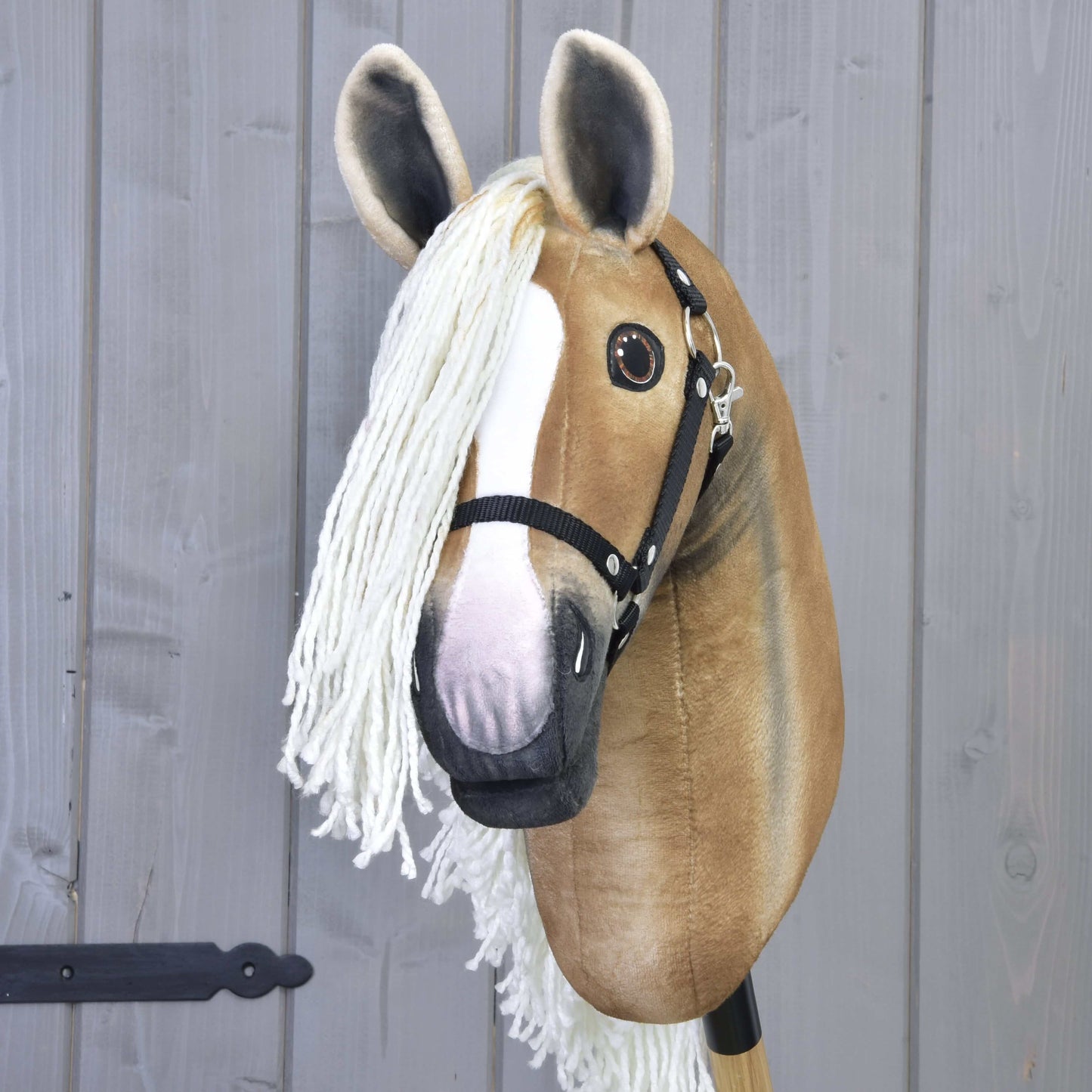 Hobby Horse Connie s černou ohlávkou od Hobby Horse LarDen s ručně vyrobeným béžovo-bílým obličejem, velkýma očima, dlouhou bílou přízovou hřívou a černou ohlávkou, nasazený na tyči před šedou dřevěnou stěnou.