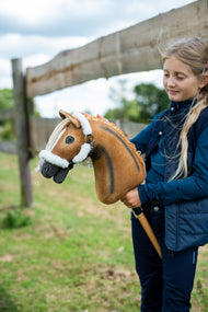 Dievča v tmavomodrej veste stojí pri drevenom ohradníku a drží Hobby Horse Connie with Black Halter od Hobby Horse LarDen s gaštanovohnedou plyšovou hlavou s tmavým chrbtovým pruhom a krémovou priadzovou hrivou, s ohlávkou Teddy Classic s kožušinovou podšívkou
