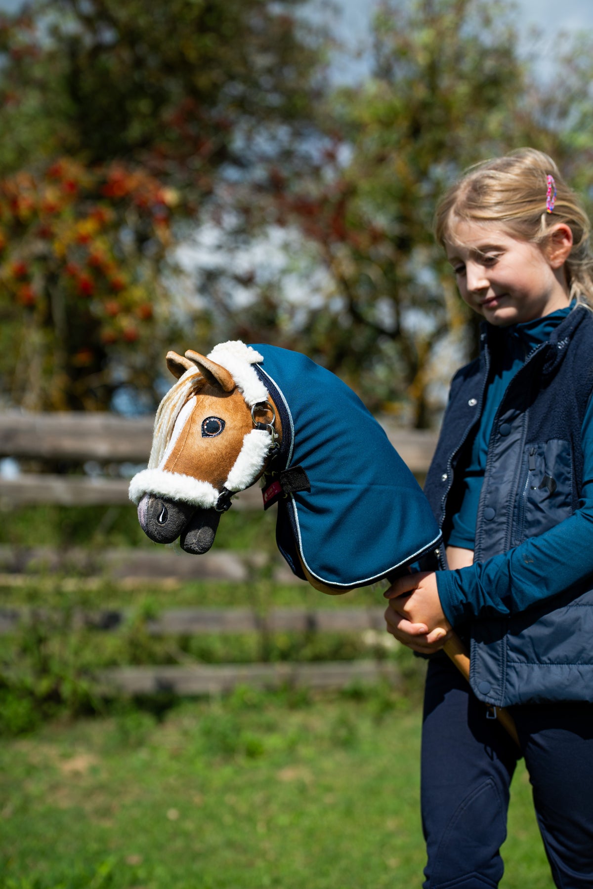 Dievča v tmavomodrej veste drží Hobby Horse Connie with Black Halter od Hobby Horse LarDen v petrolejovomodrej stajňovej deke, v pozadí sú jesenné bobule, s gaštanovohnedou plyšovou hlavou a krémovou priadzovou hrivou, s ohlávkou Teddy Classic s kožušinovou podšívkou a stajňovou dekou Winter Sky