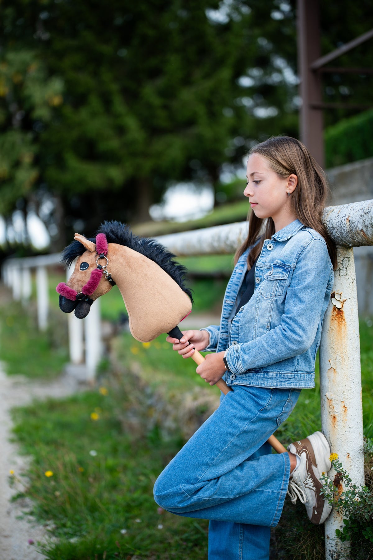 Dievča v džínsoch sa opiera o biely ohradník a drží Hobby Horse Cookie with Black Halter od Hobby Horse LarDen s pieskovou plyšovou hlavou a čiernou priadzovou hrivou, s ohlávkou Teddy Maroon s kožušinovou podšívkou