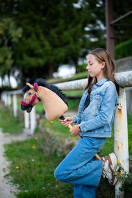 Dievča v džínsoch sa opiera o biely ohradník a drží Hobby Horse Cookie with Black Halter od Hobby Horse LarDen s pieskovou plyšovou hlavou a čiernou priadzovou hrivou, s ohlávkou Teddy Maroon s kožušinovou podšívkou