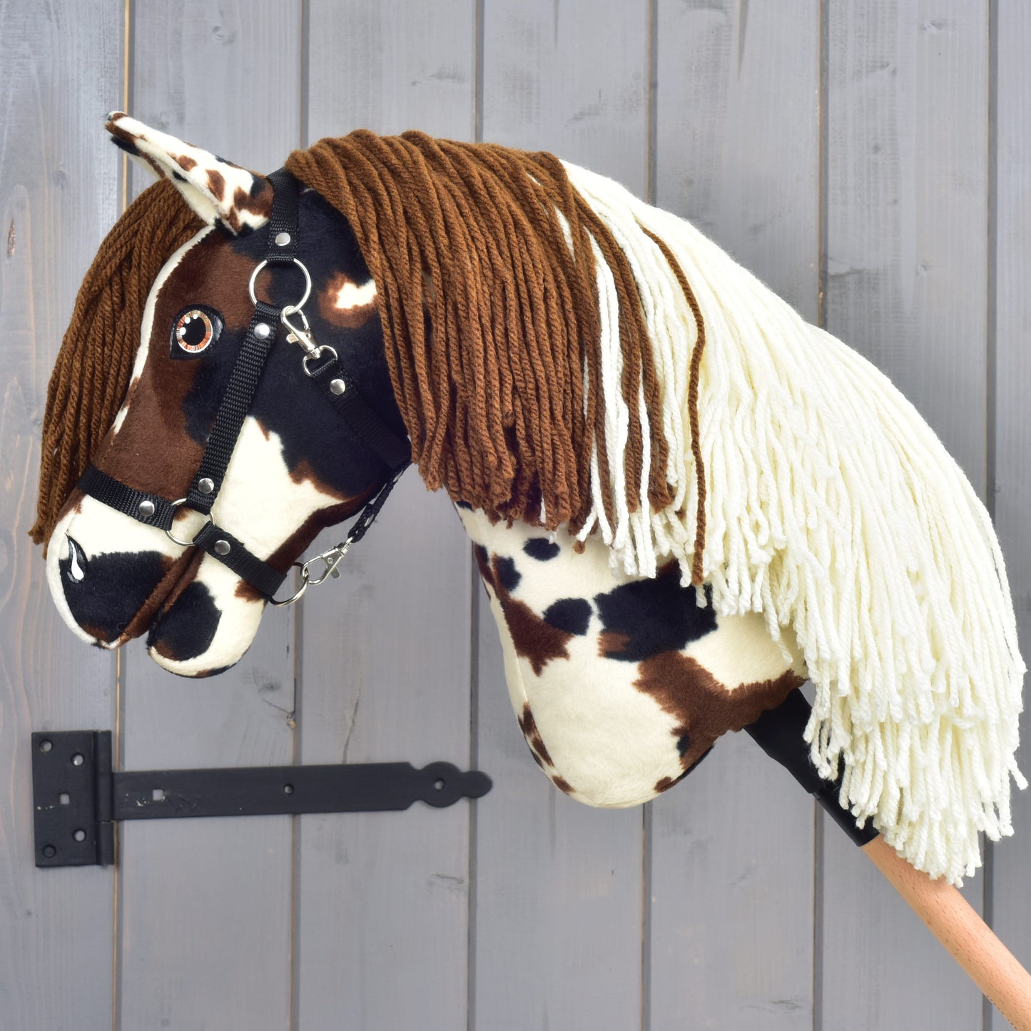 Hobby Horse Dakota s čiernou ohlávkou od Hobby Horse LarDen, ručne vyrobený model inšpirovaný divokým duchom legendárnych voľne žijúcich koní Severnej Ameriky. Má výrazné hnedé a biele škvrny, mäkkú hnedú priadzovú hrivu, krémovú priadzovú ofinu a je upevnený na drevenej tyči; stojí pred sivou drevenou stenou ako pocta neskrotnému mustangovi.
