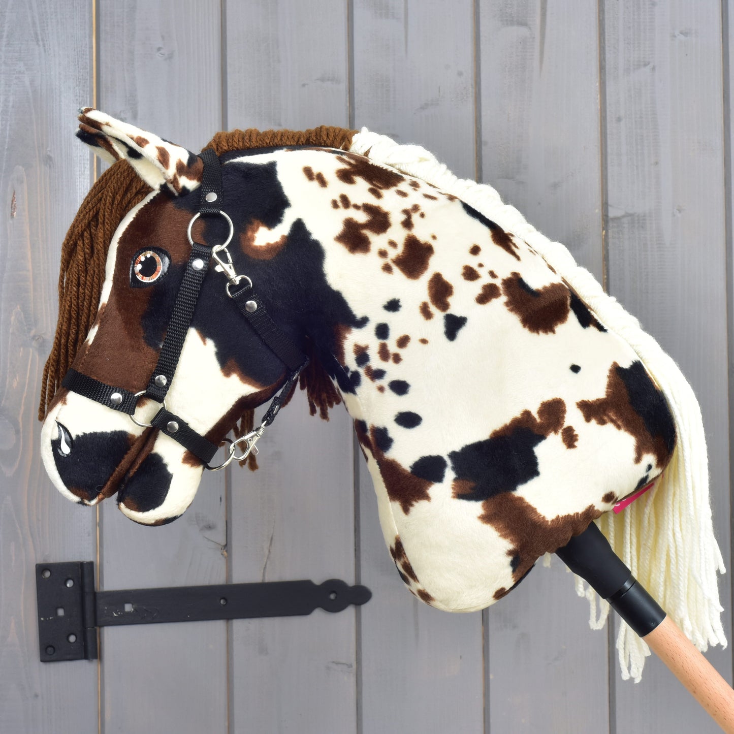 Hobby Horse Dakota s čiernou ohlávkou od Hobby Horse LarDen, plyšový koník na tyči s hnedými a bielymi škvrnami, uzdečkou a priadzovou hrivou inšpirovaný divokými mustangmi Severnej Ameriky, zobrazený na pozadí sivej drevenej steny.