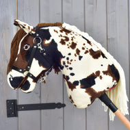 Hobby Horse Dakota s černou ohlávkou od Hobby Horse LarDen, plyšový koník na tyči s hnědými a bílými skvrnami, uzdečkou a přízovou hřívou inspirovaný divokými mustangy Severní Ameriky, zobrazený na pozadí šedé dřevěné stěny.