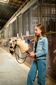 Dievča v džínsovej bunde vedie Hobby Horse Dancing Lady with Black Halter od Hobby Horse LarDen uličkou stajne s krémovou plyšovou hlavou, svetlou priadzovou hrivou a ofinou, s čiernou koženkovou ohlávkou s kompletnou čiernou kožušinou a čiernymi opratami