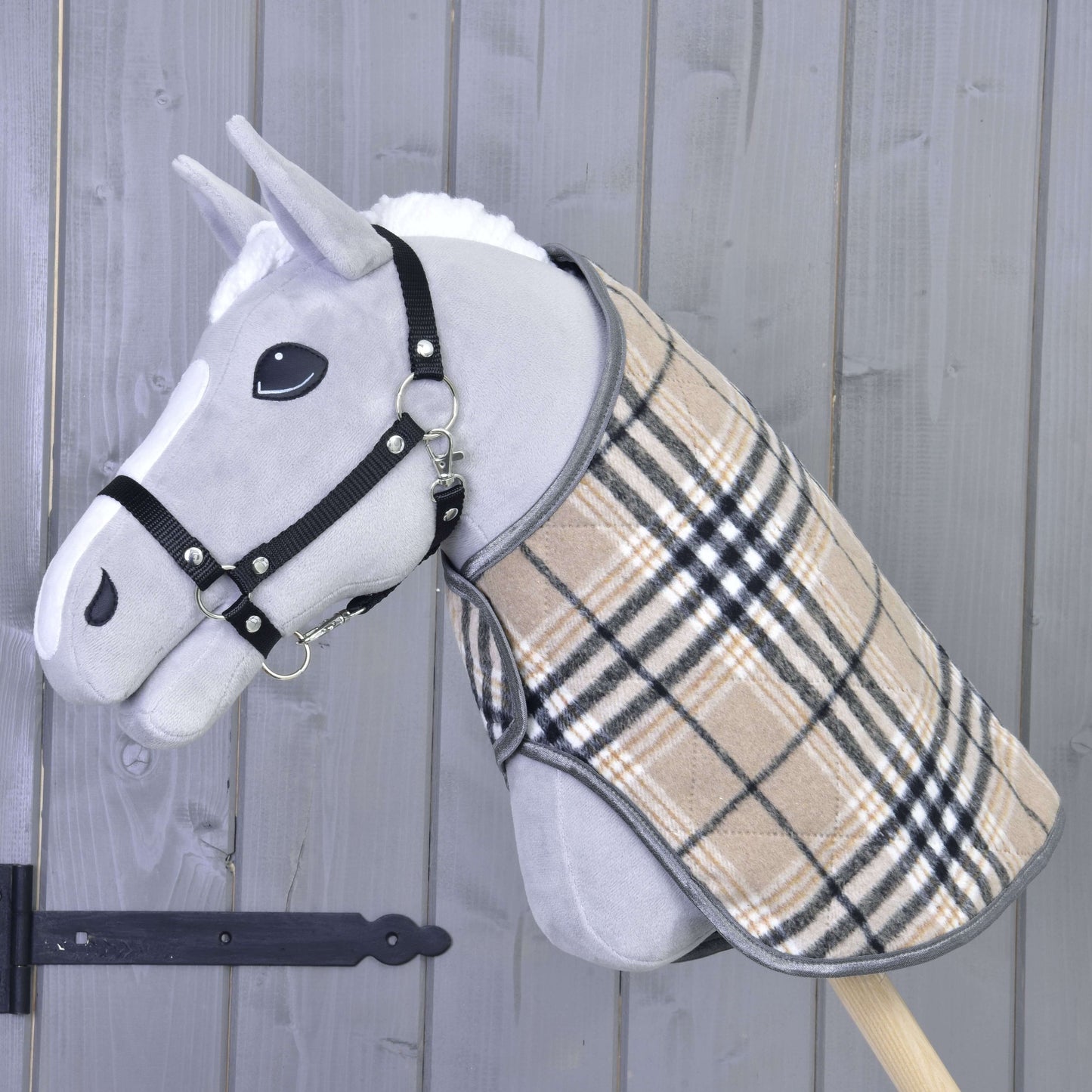 Hobby Horse LarDen „Deka Cremo England“ na sivom Hobby Horse s čiernou ohlávkou a béžovým károvaným kabátikom, doplnená čabrakou na ochranu pred hmyzom, nasadenom na drevenej tyči pred sivou drevenou stenou.