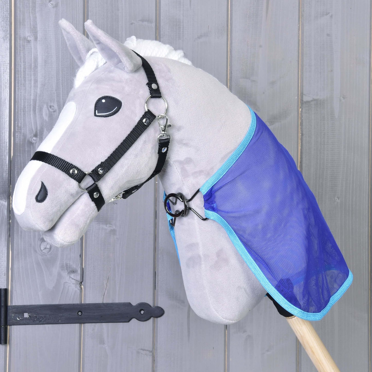 Sivá hračka Hobby Horse LarDen s čiernou uzdečkou a bielou hrivou má na sebe modrú Deka proti hmyzu. Kôň má oči v štýle kreslenej postavičky a je umiestnený pred drevenou stenou.
