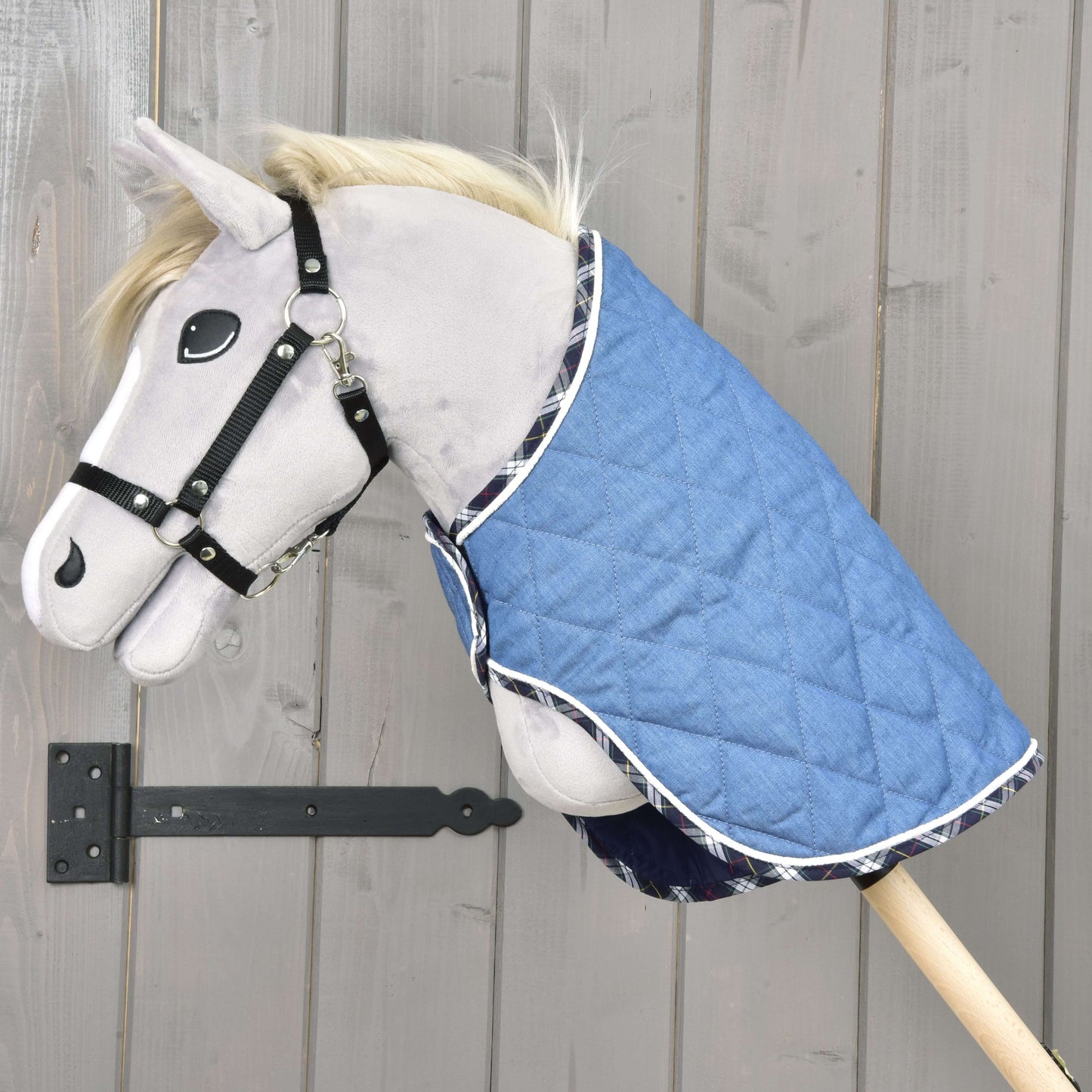 Plyšový Hobby Horse na drevenej tyči má na sebe deku Beautiful Jeans od Hobby Horse LarDen a čiernu ohlávku, umiestnený pred svetlou drevenou stenou pre dodatočnú ochranu.