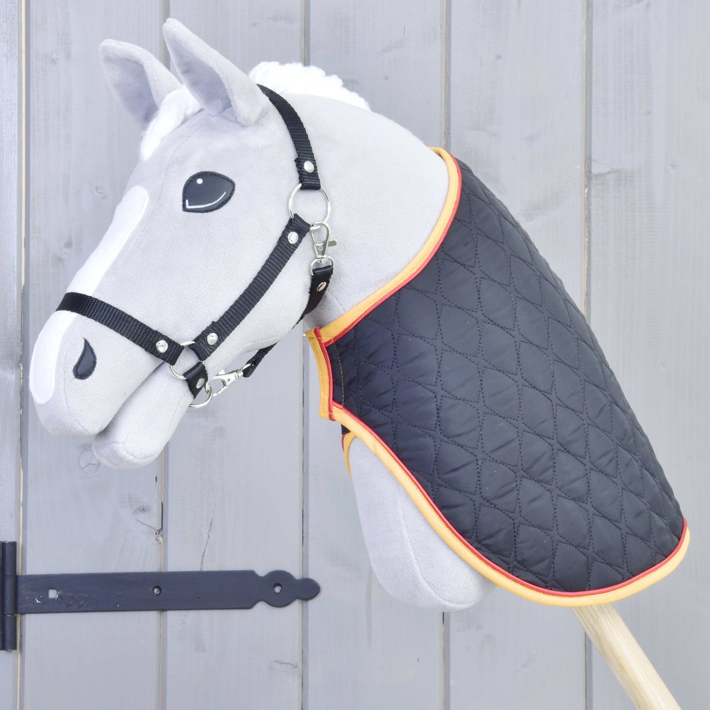 Deka Flag od Hobby Horse LarDen na sivej hračke Hobby Horse s čiernou uzdečkou a prešívanou dekou s oranžovým lemovaním, nasadenej na drevenej tyči pred sivým dreveným plotom.