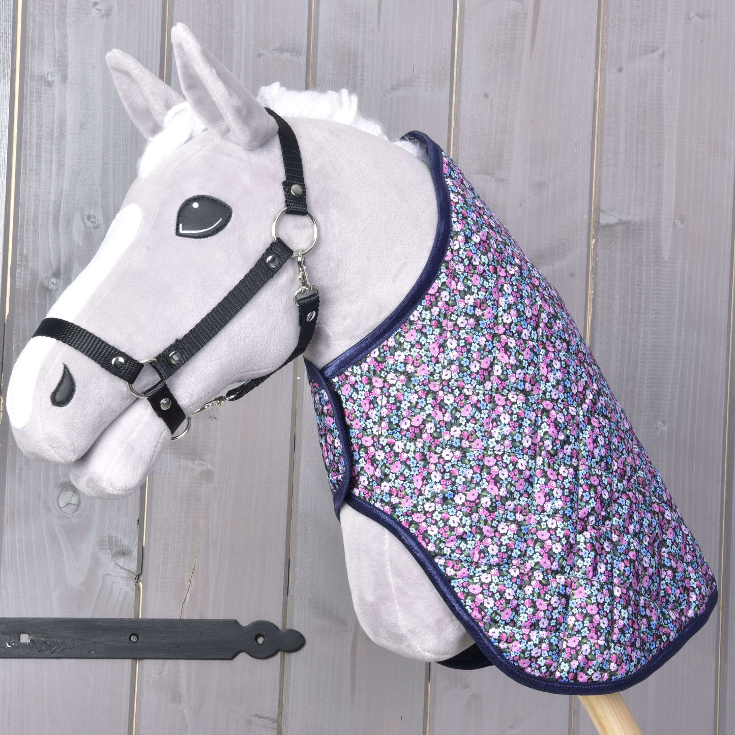Sivý Hobby Horse s bielou hrivou a čiernou uzdečkou má na sebe deku Hobby Horse LarDen Blanket Flower – farebnú deku s kvetinovým vzorom – na pozadí drevených panelov.